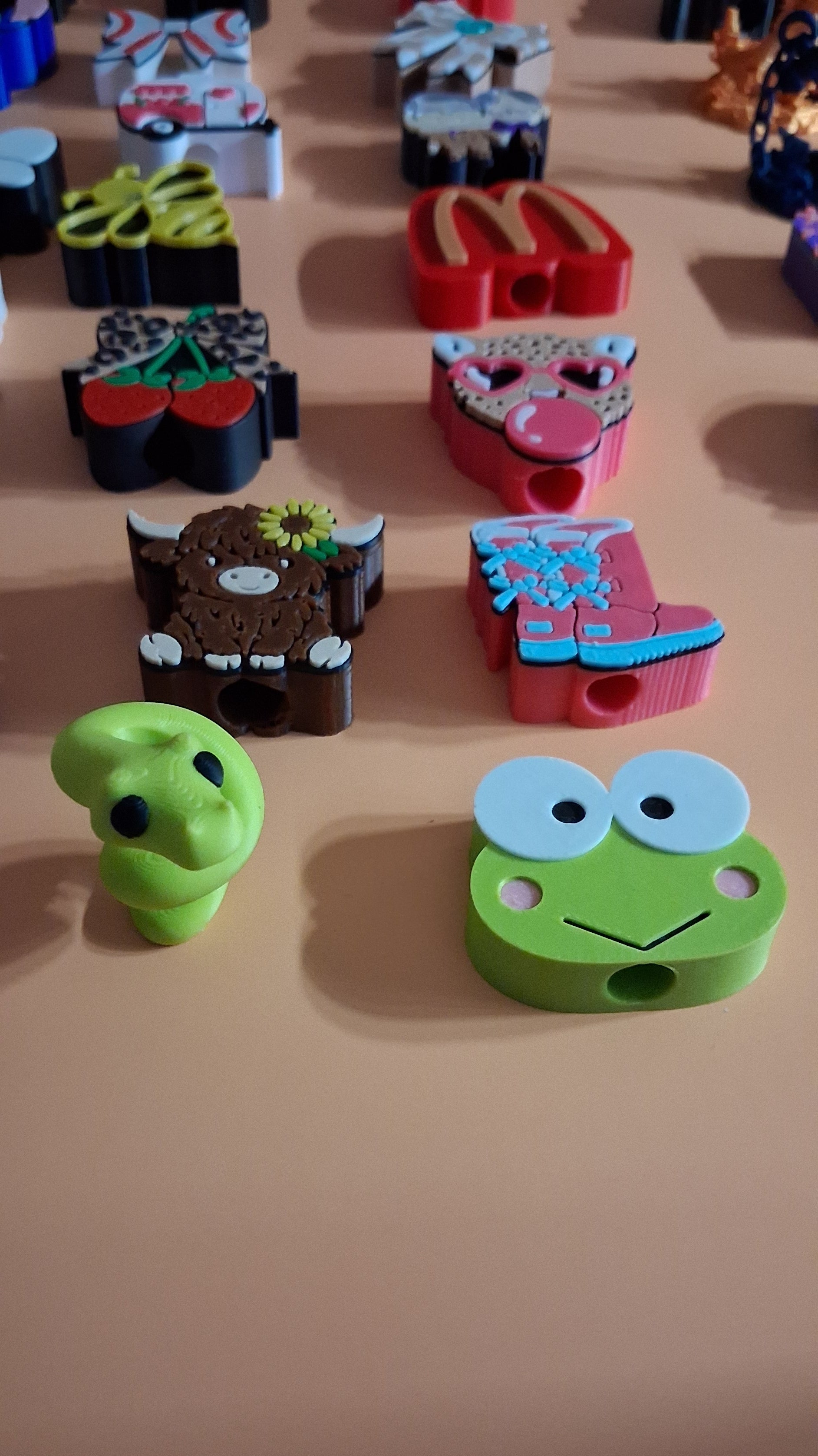 A close-up of several colorful, cartoon-style straw toppers displayed on a beige wall. The most prominent item is a green frog-shaped hook with large white eyes and a small smile, with other whimsical character hooks partially visible behind it.