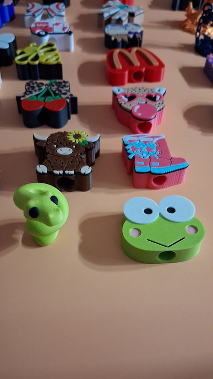 A close-up of several colorful, cartoon-style straw toppers displayed on a beige wall. The most prominent item is a green frog-shaped hook with large white eyes and a small smile, with other whimsical character hooks partially visible behind it.