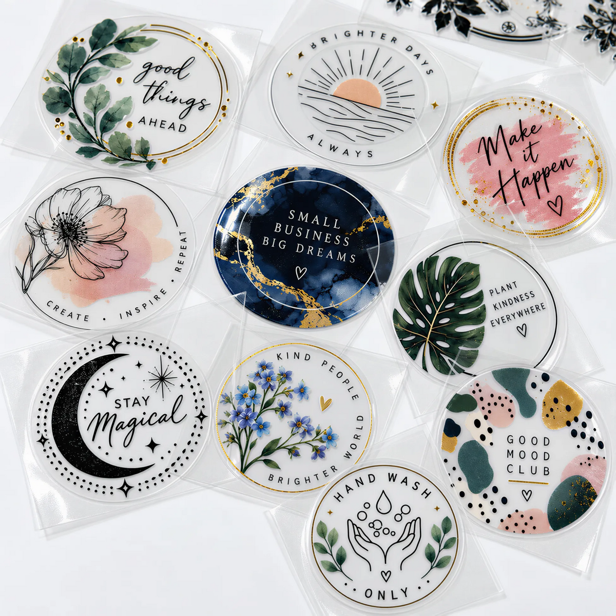 A set of round UV DTF Stickers in clear plastic sleeves, each featuring a different colorful graphic design with motivational quotes, botanical illustrations, and abstract patterns.